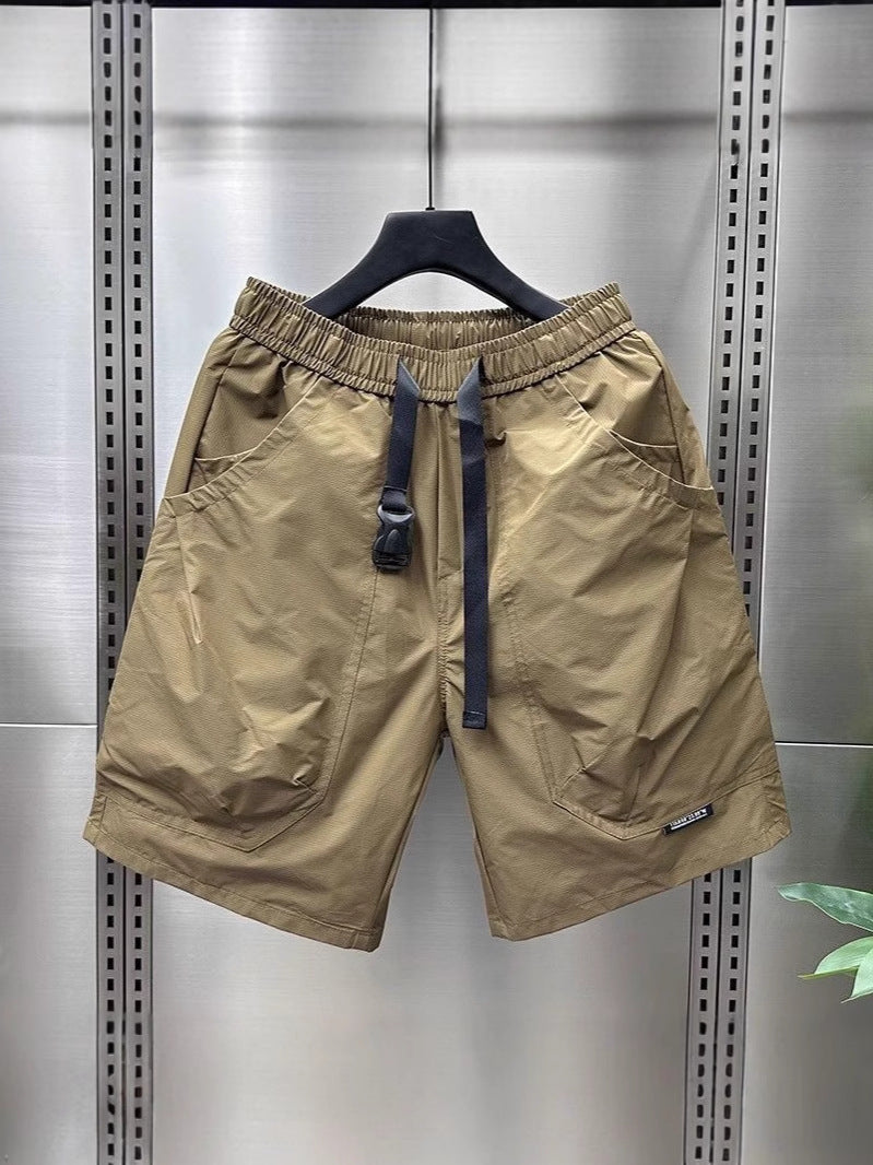 Khaki/tan cargo shorts with multiple pockets, black drawstring waist, hanging on a black hanger against a metallic background.