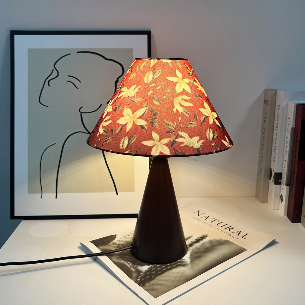Medieval style table lamp with orange floral pattern shade and conical metal base, lit on a tabletop with books and art.