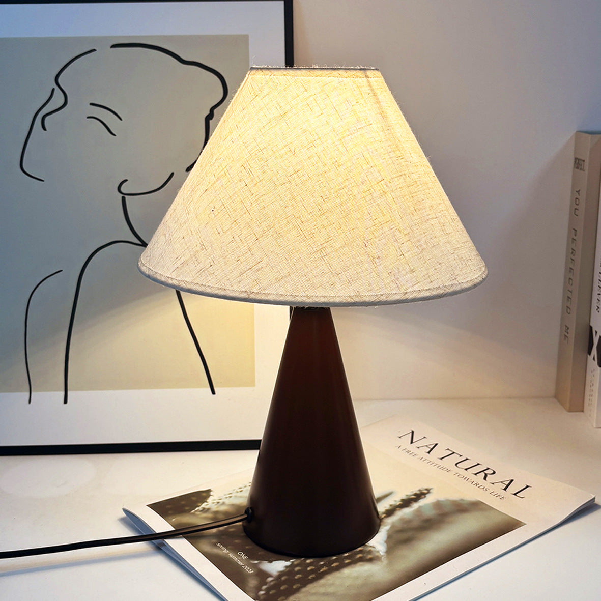 Medieval table lamp with plain beige linen shade and dark conical metal base on a desk with framed art.