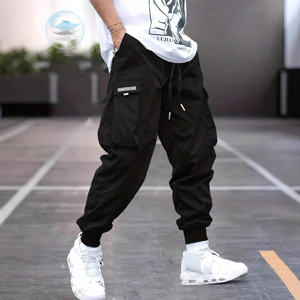Person standing on concrete pavement wearing loose black cargo jogger pants with large flap pockets, drawstring waist, elastic cuffed ankles, and white chunky sneakers. Paired with a white graphic T-shirt.