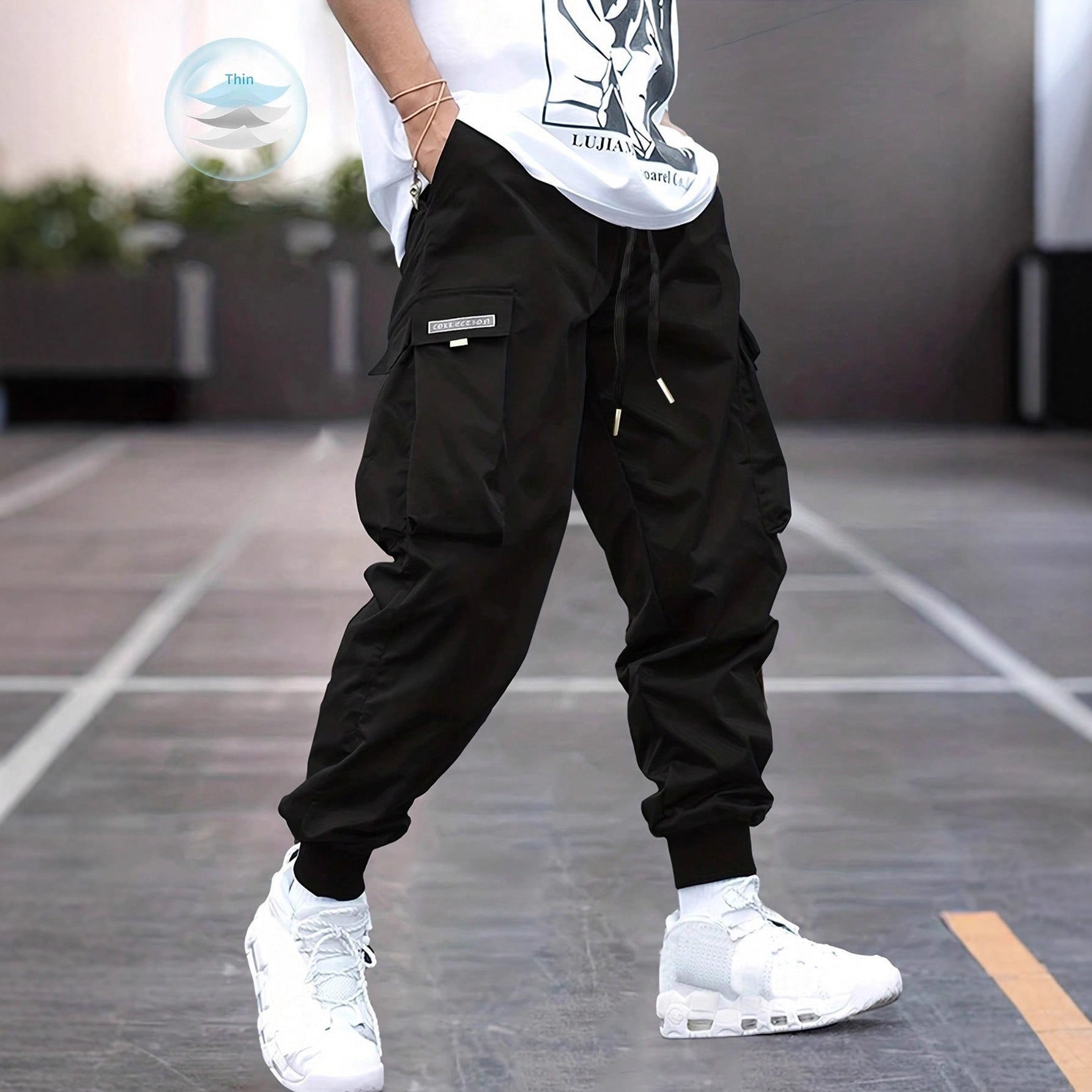 Person standing on concrete pavement wearing loose black cargo jogger pants with large flap pockets, drawstring waist, elastic cuffed ankles, and white chunky sneakers. Paired with a white graphic T-shirt.